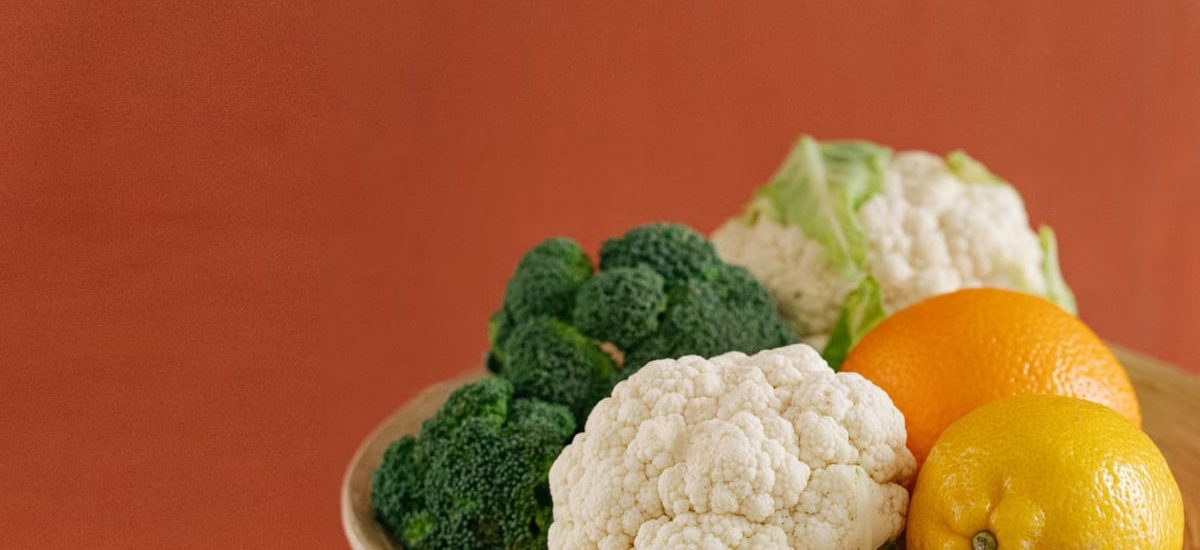 Close-up of fresh broccoli, cauliflower, lemon, and orange on a wooden plate with an earthy background.