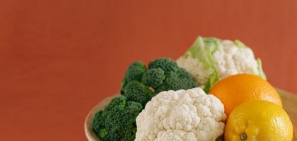 Close-up of fresh broccoli, cauliflower, lemon, and orange on a wooden plate with an earthy background.