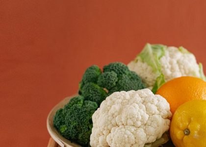 Close-up of fresh broccoli, cauliflower, lemon, and orange on a wooden plate with an earthy background.